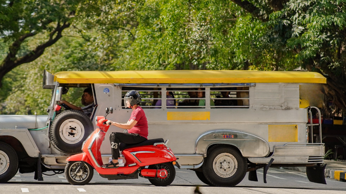 Red scooter riding beside a jeepney on a tree-lined street in the Philippines